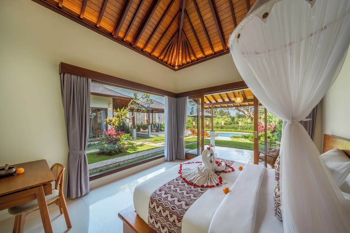 A bedroom with a bed, a dining table, and chairs. There is a view of a garden and a pool outside the window.