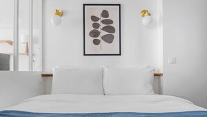 A bedroom with a bed, white bedding, a blue blanket, two wall-mounted lamps, and a framed abstract artwork. - Blueground | Le Marais, view, nr Place des Vosges (Paris)
