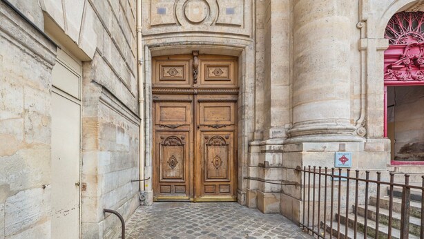 A stone building with a large wooden double door, a metal fence, and a tiled ground. - Blueground | Le Marais, view, nr Place des Vosges (Paris)
