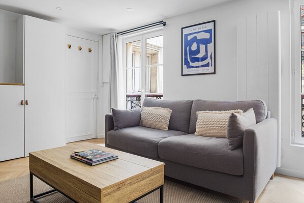 A modern living room with a grey sofa, a wooden coffee table with books, a framed artwork on the wall, and a window with a view.