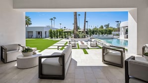 A modern outdoor lounge area with seating, a pool, and a view of a well-maintained lawn and palm trees. - SWSS Premier 10BR Estate | Pool with Swim‑Up Bar | Golf Simulator | Sport Court | Sauna & Cold Pl... (Scottsdale)
