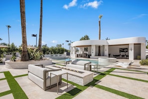 A modern outdoor pool area with lounge furniture, palm trees, and a covered seating area.