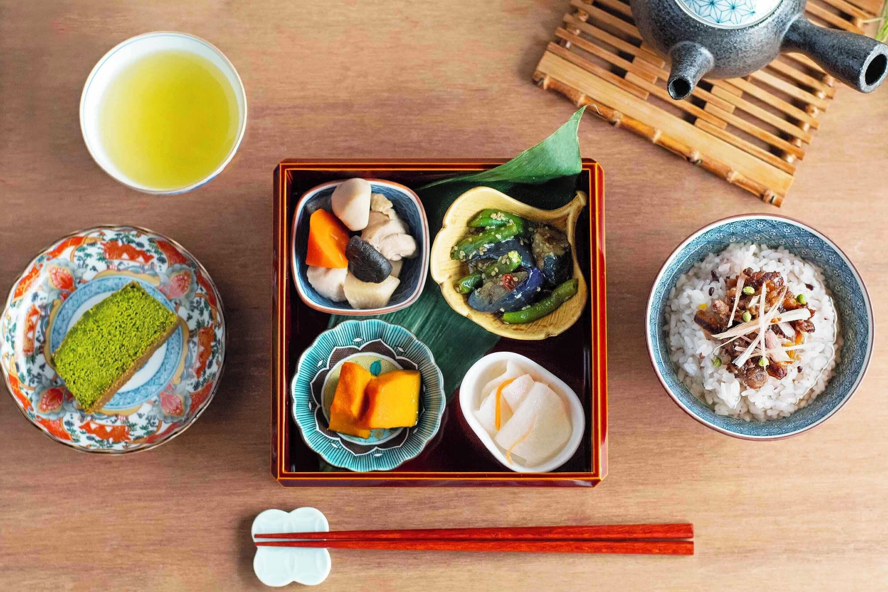 Eine traditionelle japanische Mahlzeit mit verschiedenen Gerichten, darunter Reis, Gemüse und ein grünes Dessert, serviert auf einem Holztisch mit Teekanne und Stäbchen.