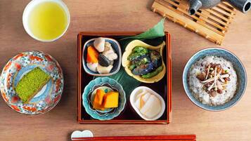 Eine traditionelle japanische Mahlzeit mit verschiedenen Gerichten, darunter Reis, Gemüse und ein grünes Dessert, serviert auf einem Holztisch mit Teekanne und Stäbchen.