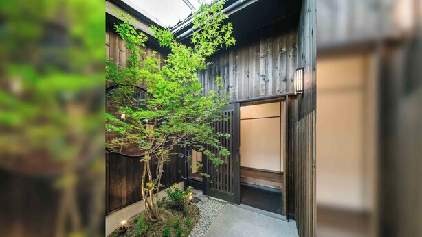 Een traditioneel Japans gebouw met een houten gevel, een kleine tuin met een boom en een betegeld pad.