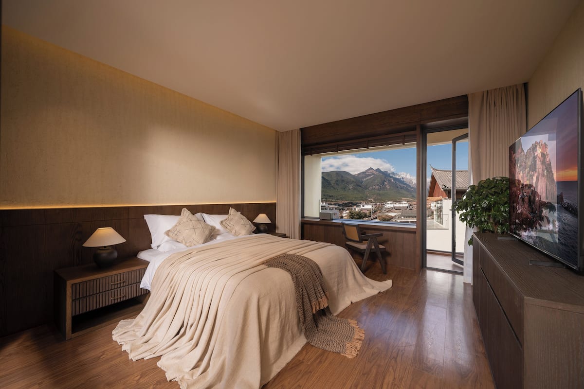 A hotel room with a large bed, a nightstand, a TV, and a view of mountains through a large window.