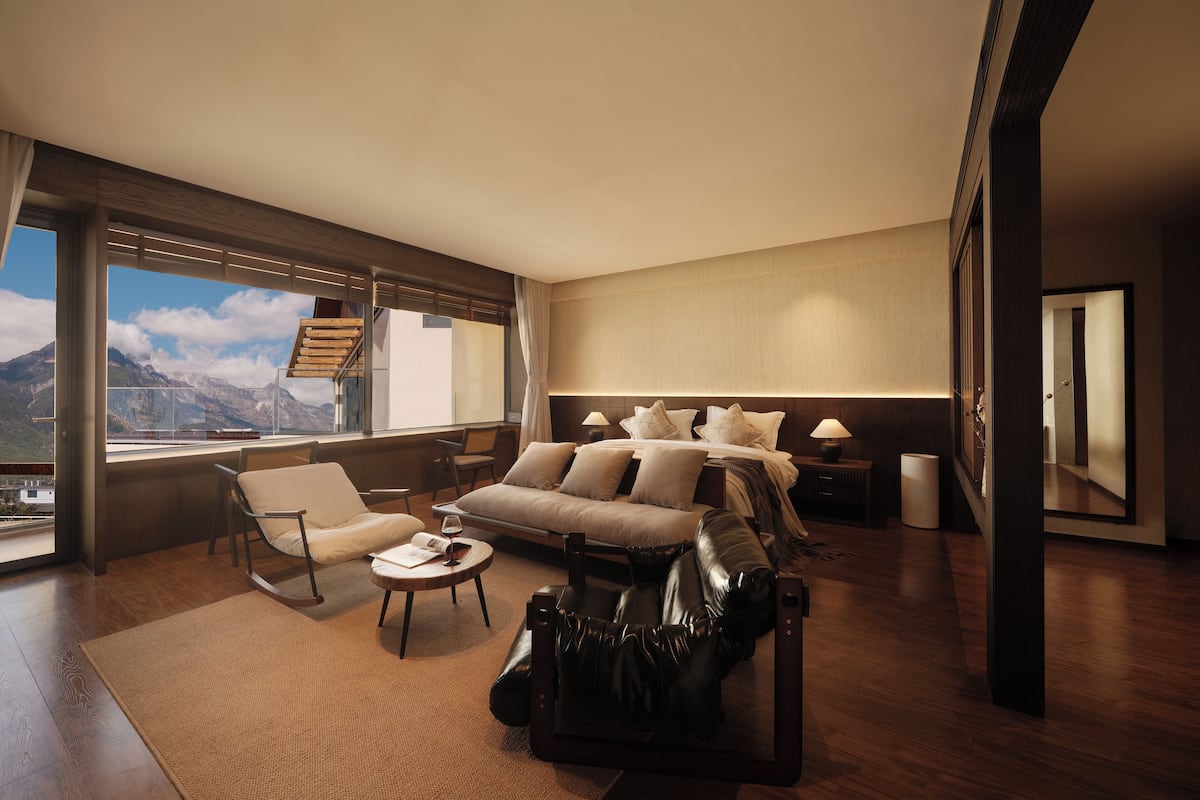 A modern hotel room with a large bed, a sofa, a coffee table, and a view of mountains through large windows.
