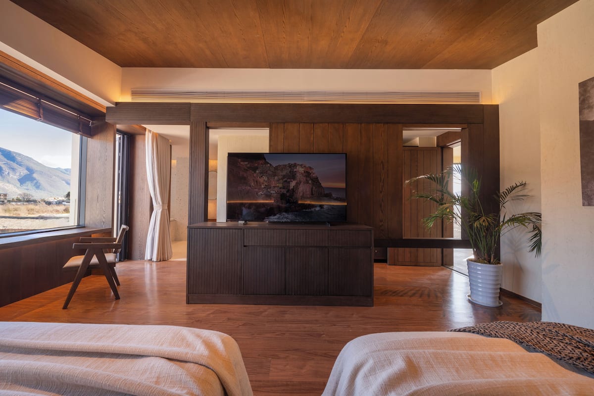 A modern hotel room with a wooden ceiling, a large flat-screen TV on a dark wooden console, a large window with a view of mountains, and a potted plant.