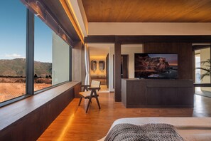Habitación de hotel moderna con un ventanal grande, suelo de madera, cama, televisor y vistas a las montañas.