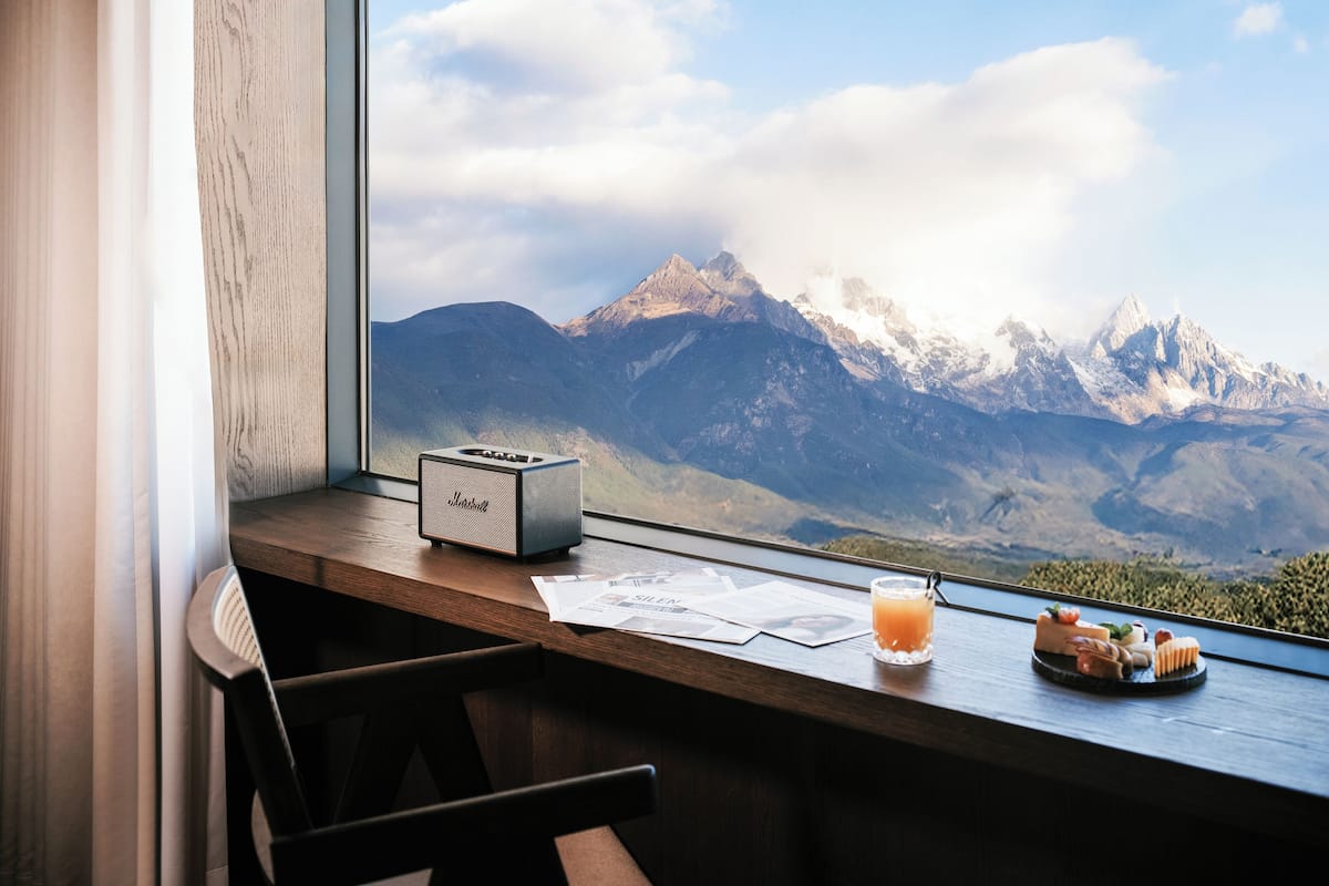 A wooden desk with a radio, papers, a glass of orange juice, and a plate of food by a window with a mountain view.