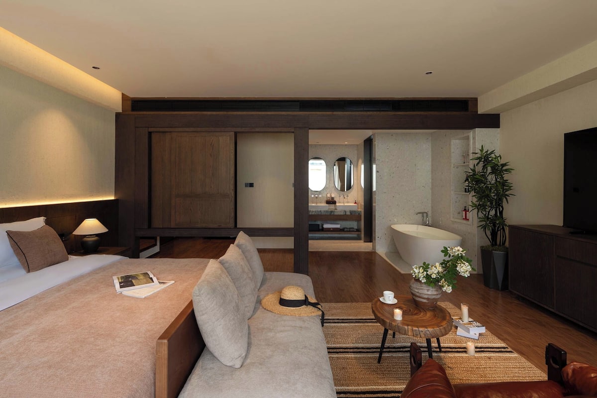 A modern hotel room with a large bed, a sofa, a small table with candles, and a view of the bathroom and kitchen.