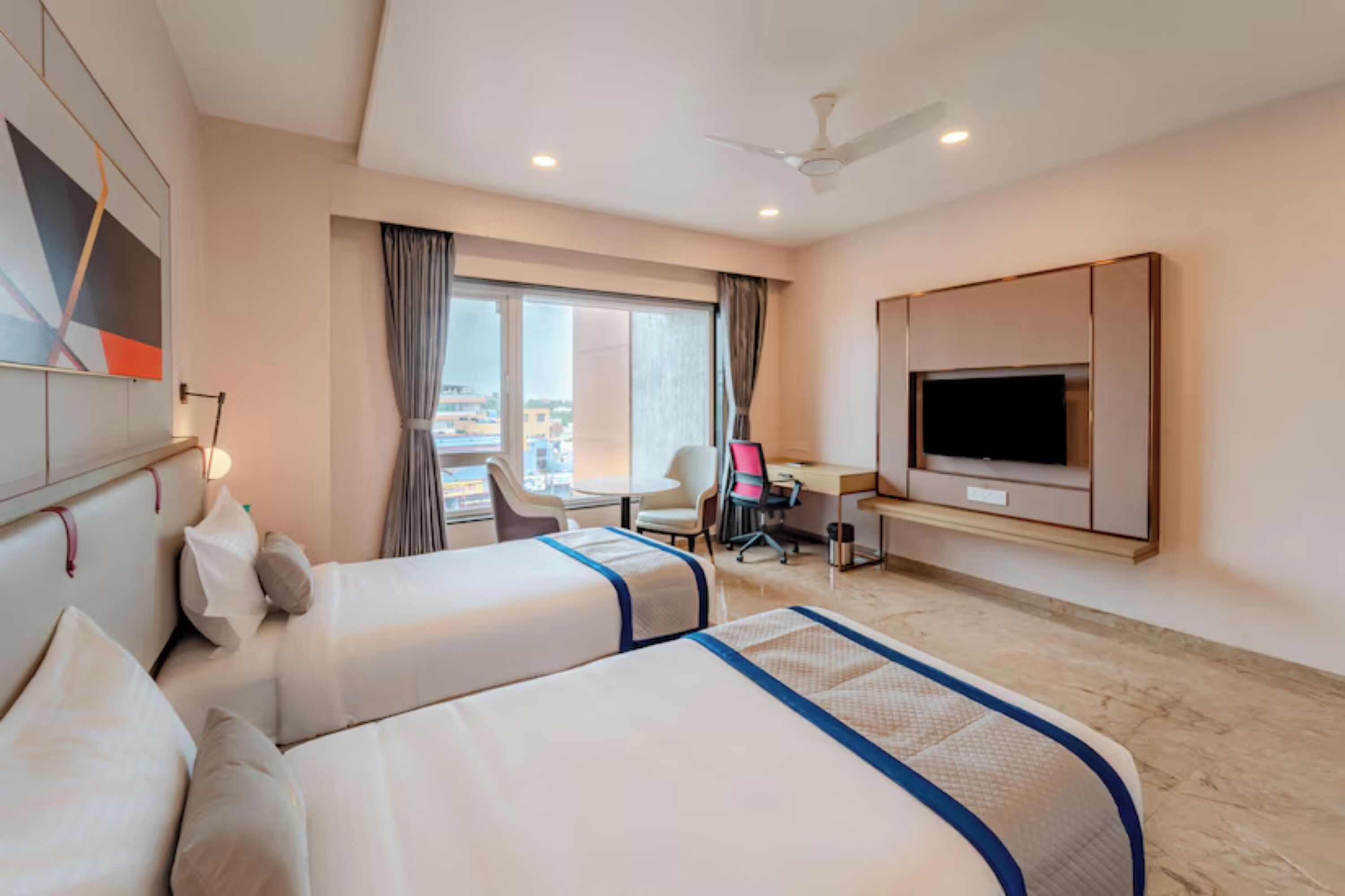 A hotel room with two beds, a desk, a chair, a TV, and a view of the city.
