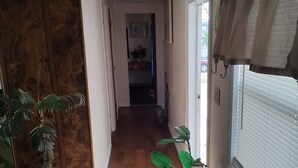 En gang med en trædør, en potteplante, et bord med et glas og et stearinlys, samt et vindue med skodder.