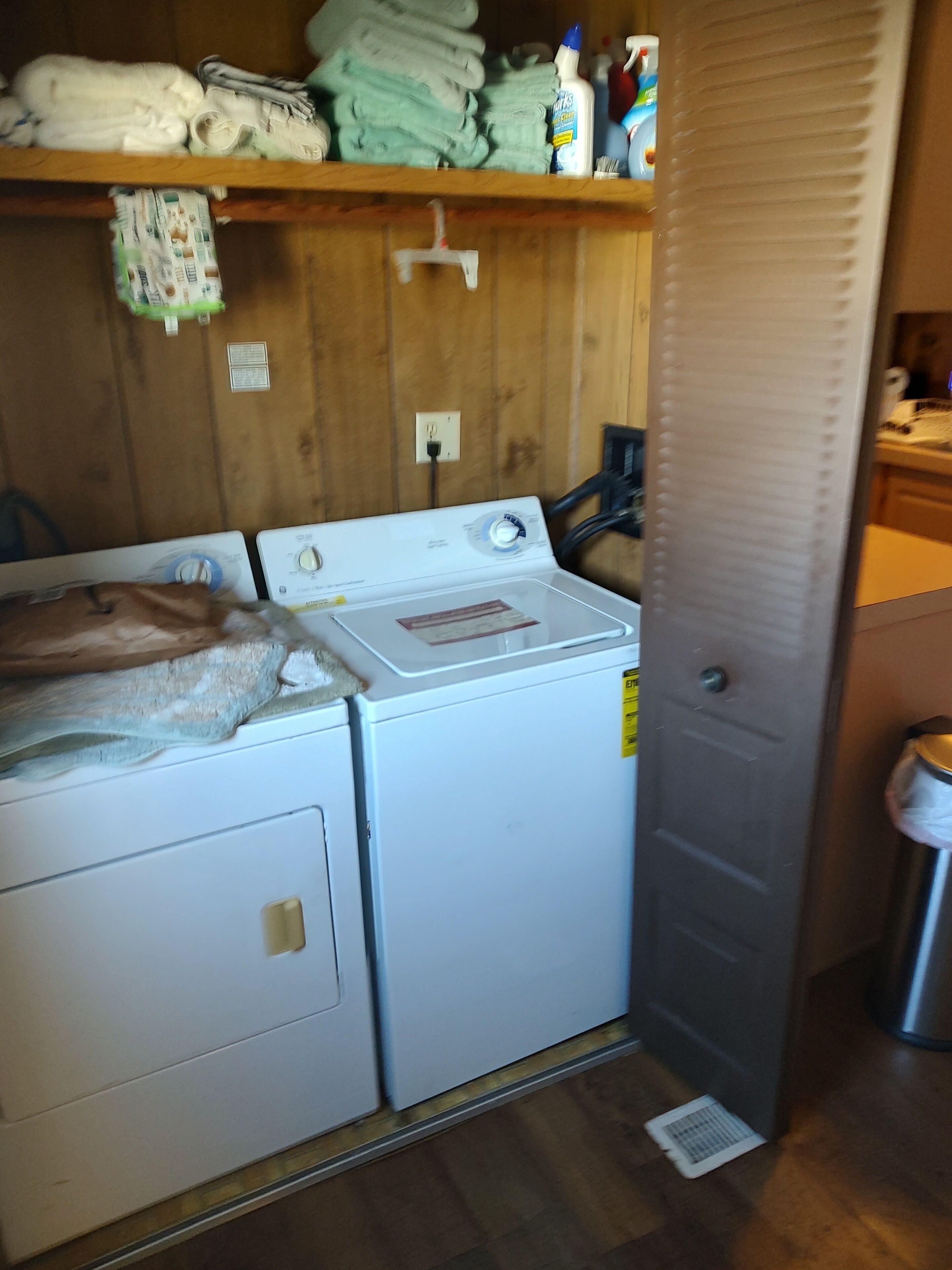 A laundry room with two stacked washers and dryers, a shelf with folded towels, and a trash can.