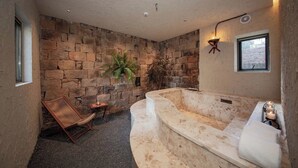 Ein Badezimmer mit einer Steinwand, einer großen Badewanne, einem hölzernen Stuhl und einem kleinen Tisch mit einer Kerze.