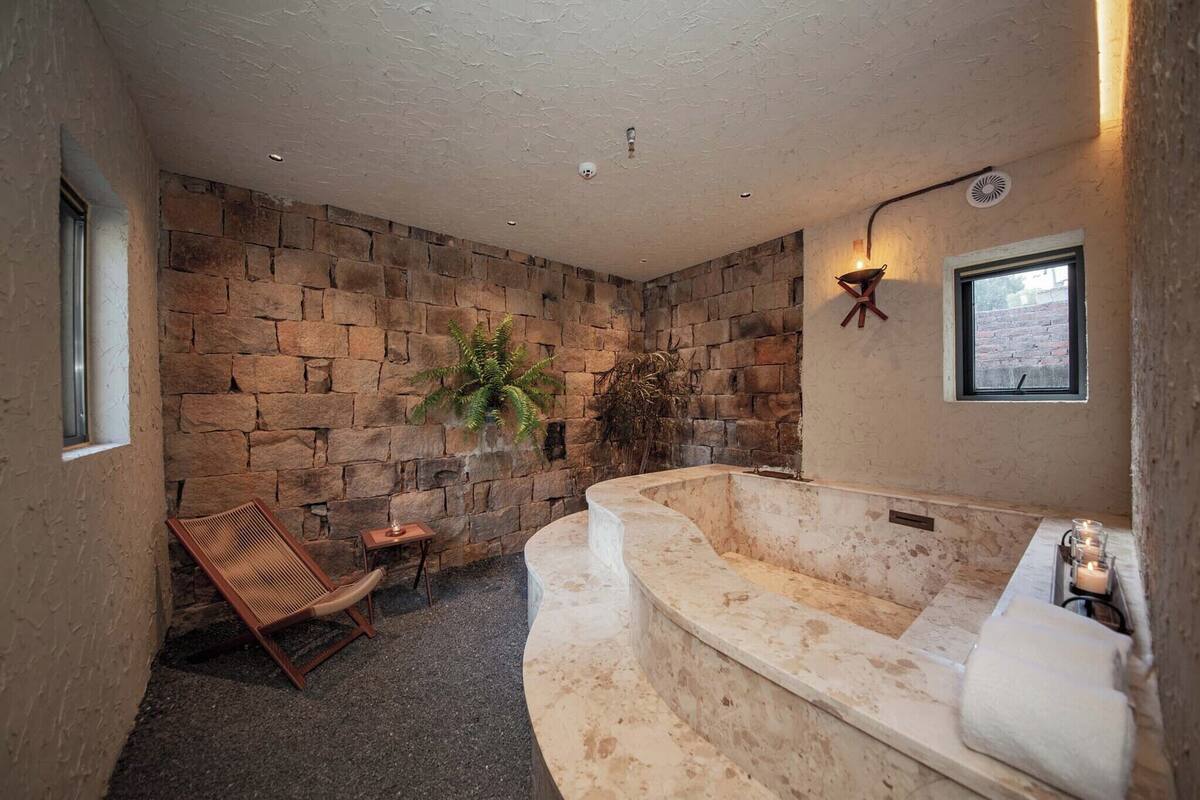 Une salle de bain avec un mur en pierre, une grande baignoire, une chaise en bois et une petite table sur laquelle est posée une bougie.
