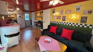 Un espace de vie compact comprenant un coin cuisine, un coin repas avec une nappe à carreaux, un canapé avec des coussins rouges et un ventilateur de plafond.
