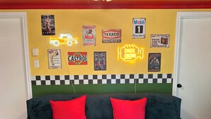 Une chambre avec une tête de lit à carreaux, un panneau néon indiquant « VÉRIFIER LE MOTEUR » et divers affiches sur le thème de l’automobile accrochées au mur.
