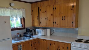 A kitchen with wooden cabinets, a white refrigerator, a microwave, a stove, and a window with a view of greenery. - Beach garden cottage with AC in charming Ogunquit (Ogunquit)
