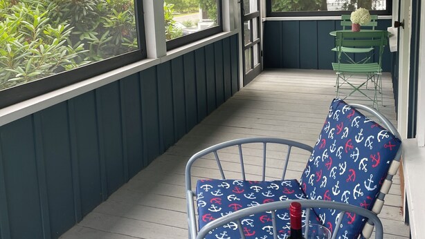 A covered porch with a table and chairs, a flag, and a view of greenery. - Beach garden cottage with AC in charming Ogunquit (Ogunquit)