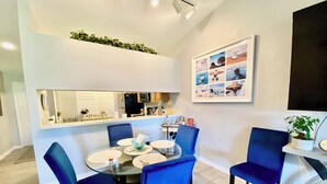 Un espace repas moderne avec une table ronde en verre, des chaises recouvertes de tissu bleu, une cuisine en arrière-plan et un collage encadré accroché au mur.