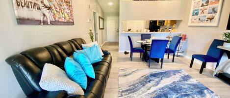 A modern living room with a black leather sofa, blue cushions, a marble-patterned rug, and a dining area with blue chairs.