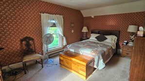 A bedroom with a bed, bedside lamps, a wooden chest, a bench, a fan, and a window with curtains.