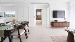 Une cuisine moderne avec des meubles blancs, un coin repas doté d’une table et de chaises en bois, et un téléviseur fixé au mur.