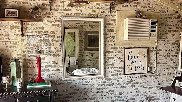 Una habitación rústica con paredes de ladrillo visto, una cama con ropa de cama floral, un espejo antiguo y un aire acondicionado de pared.