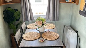 A compact dining area with a table set for four, surrounded by chairs, a radiator, and a window with curtains. - 2 Bed Caravan - Sleeps 6 - Dogs - Parking - Wifi (point)