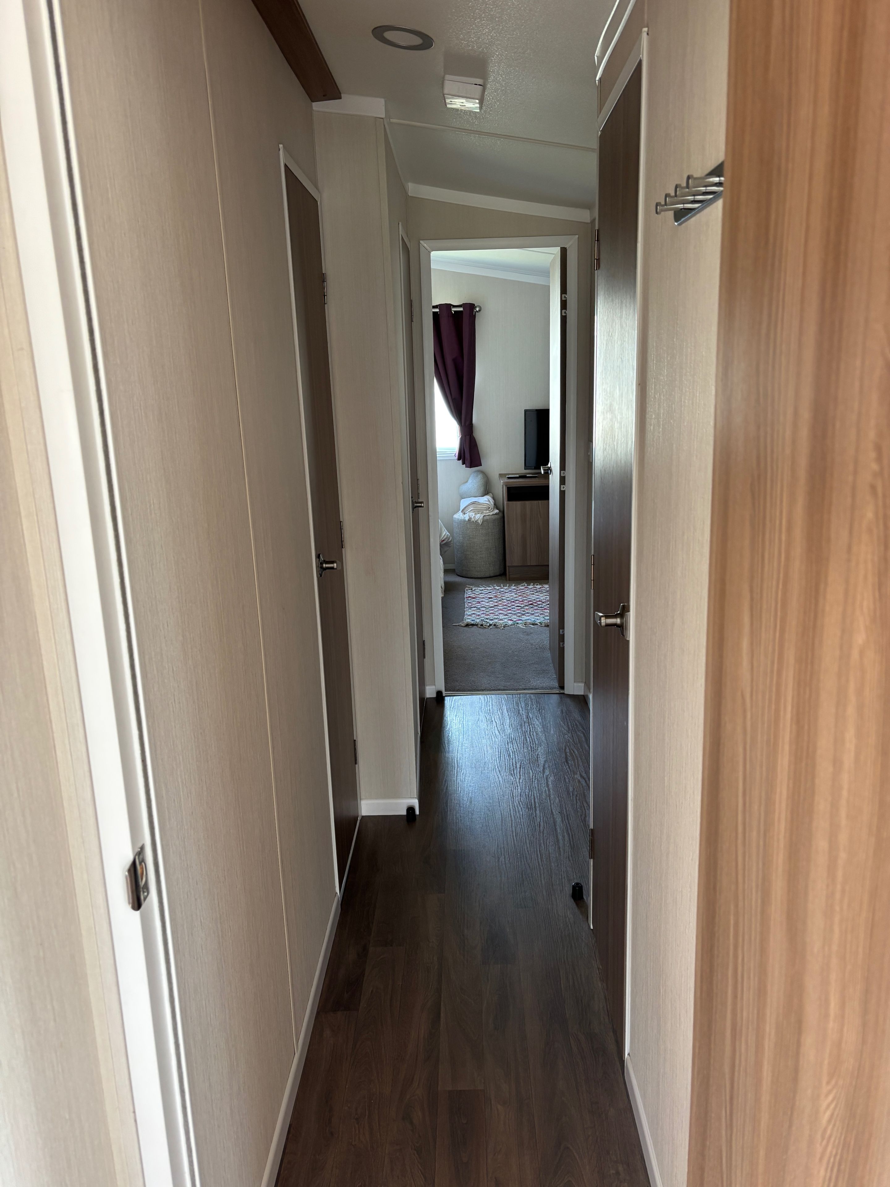 A hallway with wooden flooring leading to a bedroom with a bed, a television, and a window with curtains.