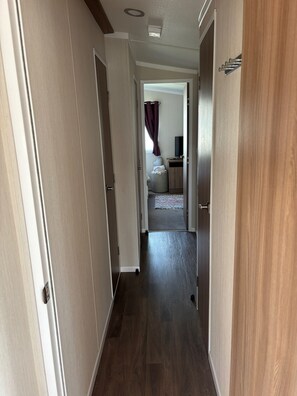 A hallway with wooden flooring leading to a bedroom with a bed, a television, and a window with curtains. - 2 Bed Caravan - Sleeps 6 - Dogs - Parking - Wifi (point)