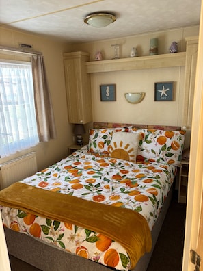 A bedroom with a bed, a window with curtains, a radiator, a shelf with decorative items, and wall art. - 2 Bed Caravan - Sleeps 6 - Dogs - Parking - Wifi (point)