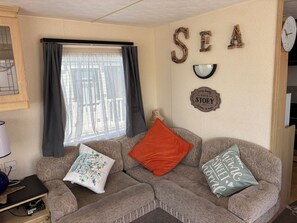 A cozy living room with a sofa, decorative pillows, a wall clock, and a window with curtains. - 2 Bed Caravan - Sleeps 6 - Dogs - Parking - Wifi (point)