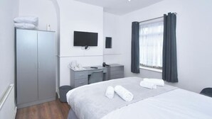 Hotellihuone, jossa on sänky, työpöytä tuolilla, vaatekaappi, seinälle asennettu televisio ja ikkuna, jossa on verhot.
