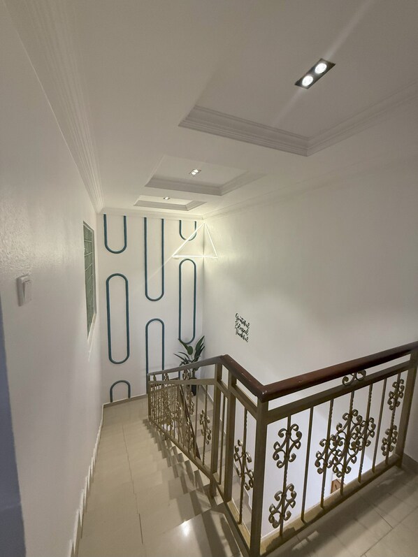 Una escalera con barandilla de madera y balaustradas metálicas decorativas, que conduce a un rellano con una lámpara fijada a la pared y una planta en maceta.