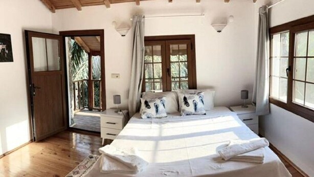 A bedroom with a large bed, wooden flooring, a window with curtains, and a door leading outside. - Spacious Stone Villa in the heart of Kayakoy, Overlooking the historic ruins. (Fethiye)