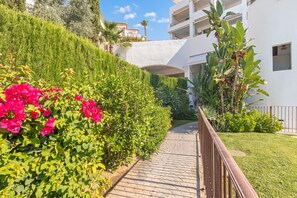A pathway leading to a building with a white facade, surrounded by lush greenery and flowering plants.