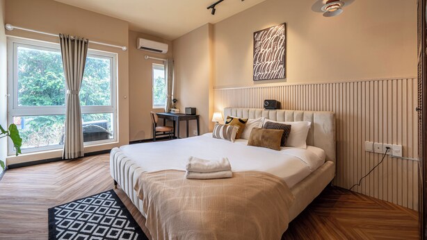 A hotel room with a large bed, a desk with a chair, a window with curtains, and a radiator on the wall.