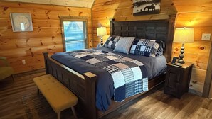 A wooden cabin bedroom with a large bed, a bench, two bedside lamps, and a window with blinds. - Modern•Prime Location•Mountain Views•Theater•Hot Tub•minutes to ALL attractions! (Sevierville)
