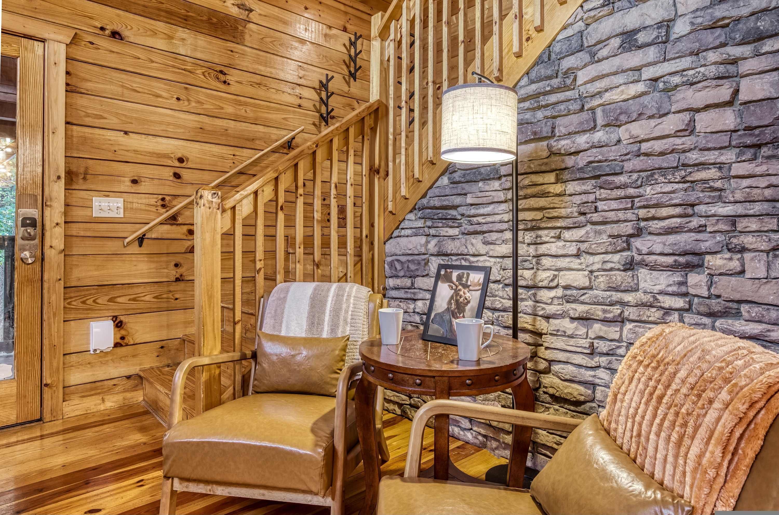 A cozy room with wooden walls, a stone wall, a staircase, leather chairs, a round wooden table, and a lamp.