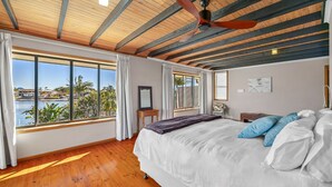 A bedroom with a large bed, wooden flooring, a ceiling fan, and a view of a body of water and buildings. - Maroochydore Waterfront (Maroochydore)
