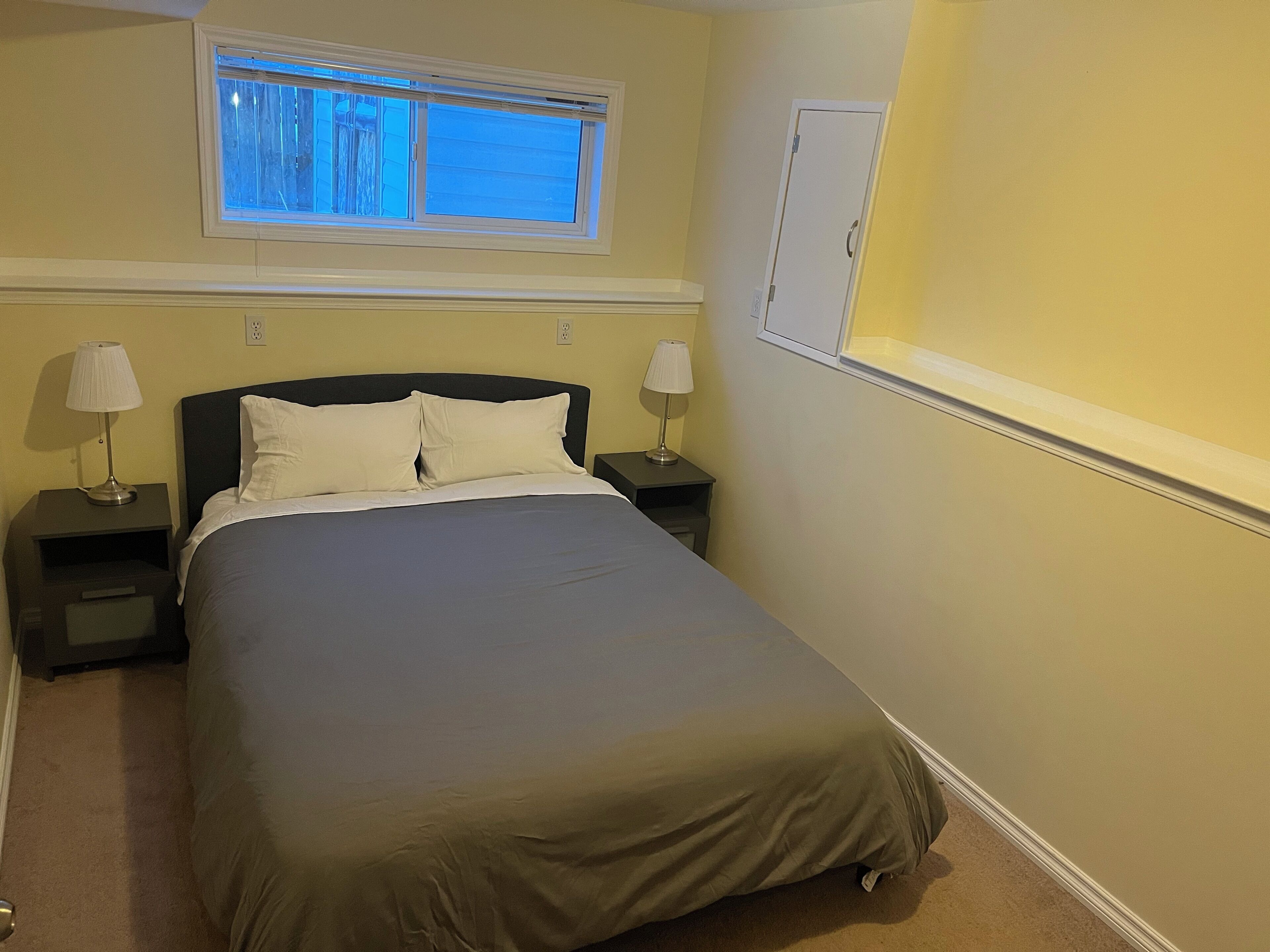 Una cama bien hecha con dos almohadas, una mesita de noche con lámpara, una ventana con persianas y una puerta.