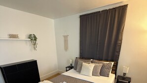 A neatly made bed with a grey quilt, a white throw blanket, and rolled towels. There is a bedside table with a lamp and a small plant. The room has wooden flooring and a dark curtain over the window.
