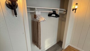 A hallway with a wooden floor, a white wall, and a coat rack with hangers and a blanket. There are two wall-mounted light fixtures on either side of the door.