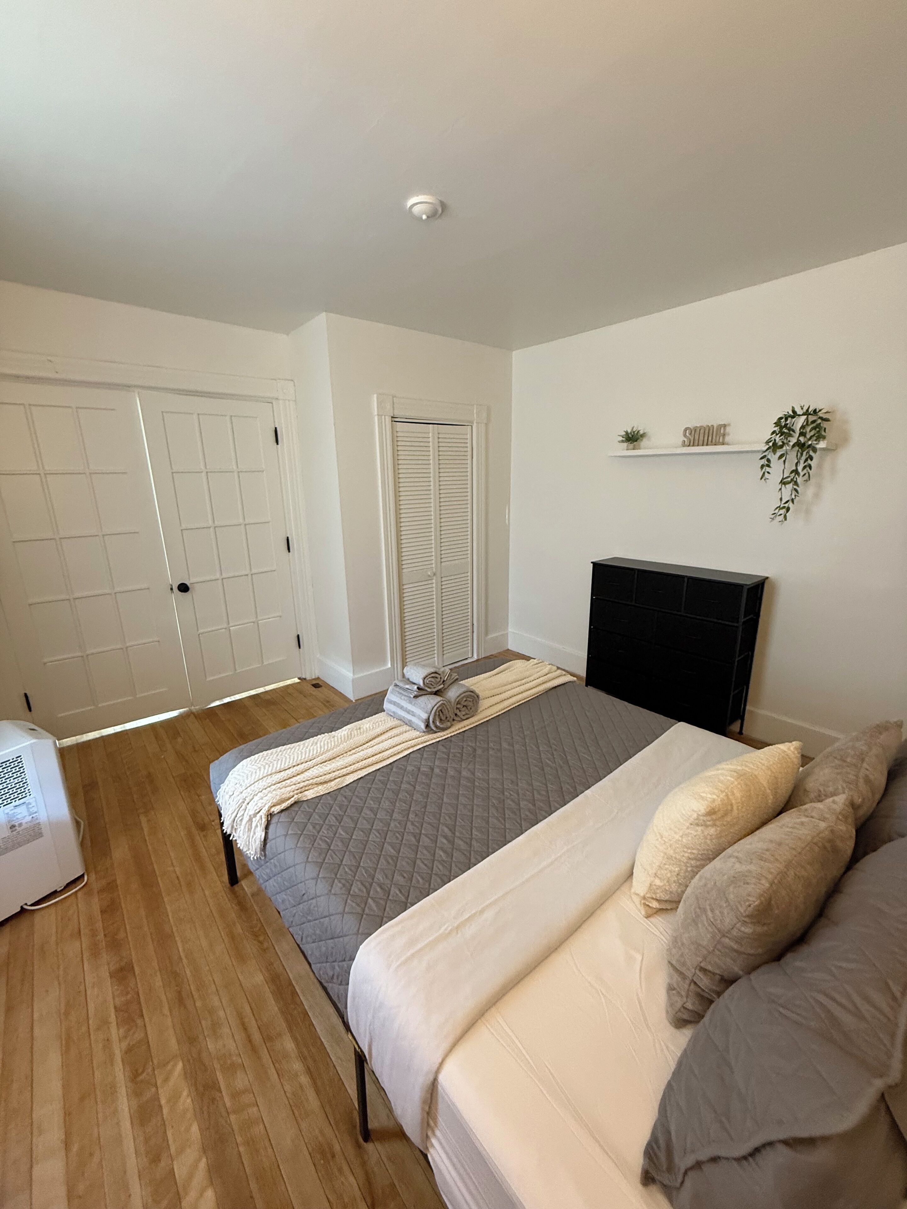 A bedroom with a bed, a bedside table, a heater, a wardrobe, and a shelf with a plant and a sign.
