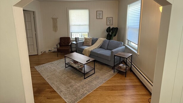 A living room with a gray sofa, a coffee table, and a rug. There are two windows with blinds, a door, and a wall-mounted plant.