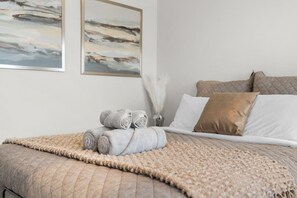 Un lit bien fait, avec des serviettes pliées, un plaid beige et un vase décoratif orné de plumes blanches.
