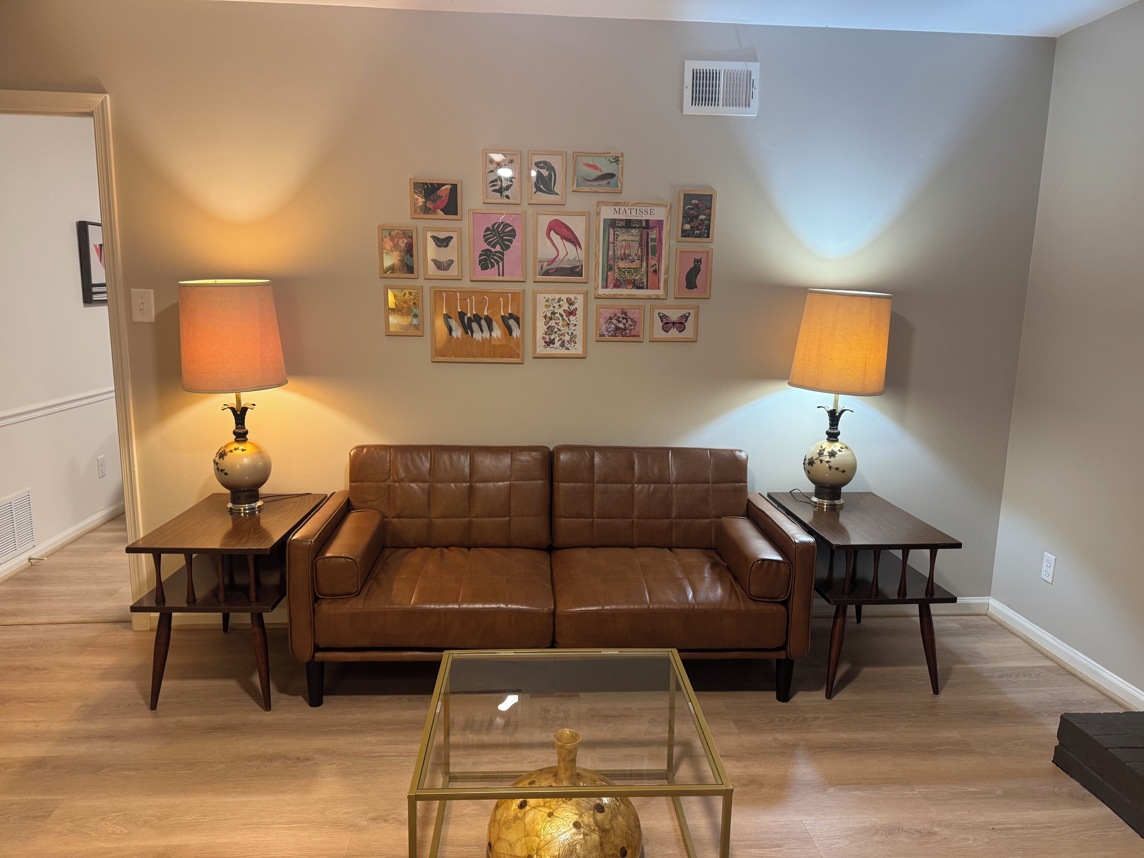 A living room with a brown leather sofa, two side tables with lamps, a glass coffee table, and a wall decorated with various framed artworks.