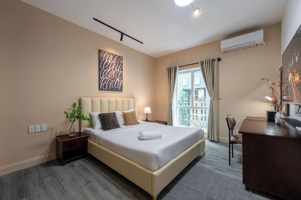 A hotel room with a large bed, a desk, a nightstand, a potted plant, and a view of the city through the window.